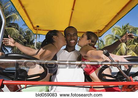 Stock Image - Hispanic women kissing African man’s cheeks. Fotosearch