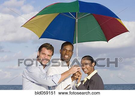 Stock Image - Multi-ethnic businesspeople under beach umbrella. Fotosearch