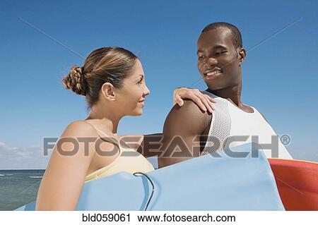 Stock Image - Multi-ethnic couple holding bodyboards. Fotosearch