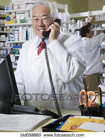 Stock Image - Senior Asian pharmacist talking on telephone. Fotosearch