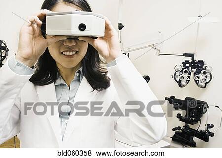 Stock Photo - Asian female optometrist looking through equipment. Fotosearch