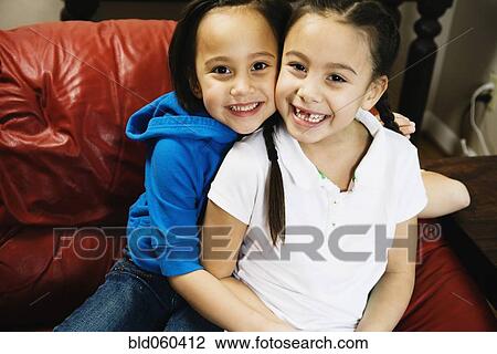 Stock Image - Asian sisters hugging on sofa. Fotosearch