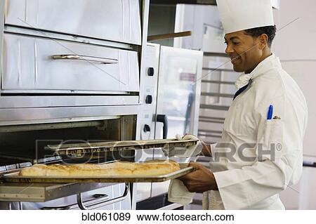 Stock Photo - African male baker taking bread from oven. Fotosearch