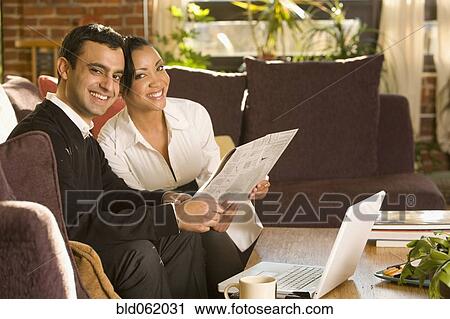 Stock Image - Mixed race couple reading newspaper in living room. Fotosearch