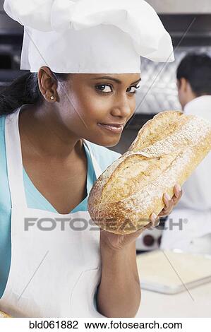 Stock Image - Mixed Race female baker smelling fresh bread. Fotosearch