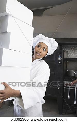 Stock Image - Mixed Race female pastry chef carrying stack of boxes. Fotosearch