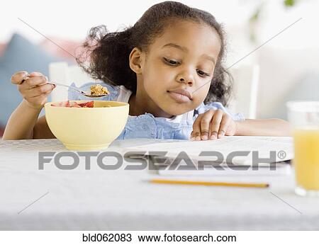 Stock Image - Mixed race girl reading and eating breakfast. Fotosearch