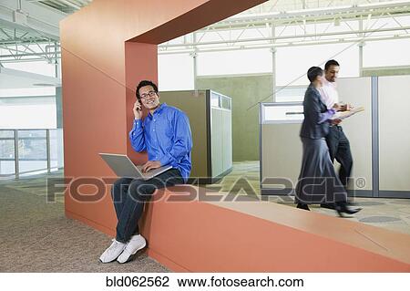 Stock Image - Asian American businessman with laptop talking on cell phone in office. Fotosearch