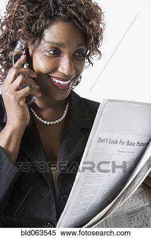 Stock Photography - Mixed race businesswoman talking on cell phone and reading newspaper. Fotosearch