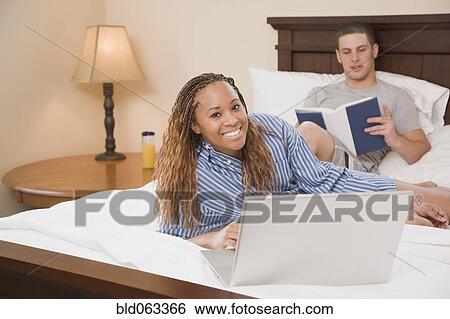 Stock Photograph - Multi-ethnic couple reading and typing on laptop in bed. Fotosearch