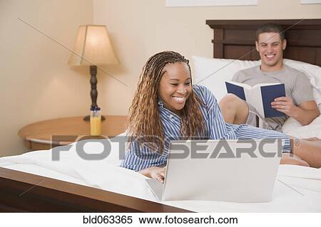 Stock Photography - Multi-ethnic couple reading and typing on laptop in bed. Fotosearch