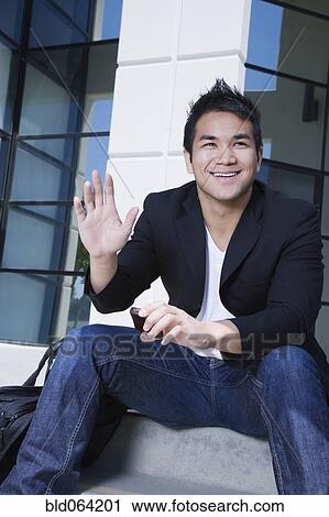 Stock Image - Asian man holding cell phone and waving. Fotosearch