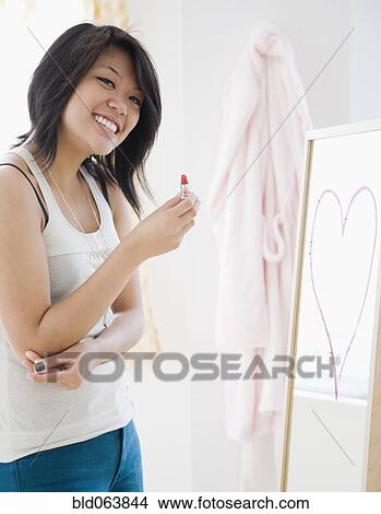 Picture - Asian woman drawing heart shape with lipstick on mirror. Fotosearch