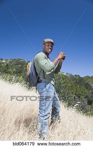 Stock Photo - Mixed race man backpacking on hillside with binoculars. Fotosearch