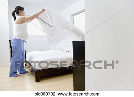 Stock Image - Pregnant Asian woman making bed. Fotosearch