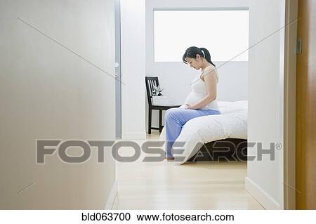 Stock Image - Pregnant Asian woman sitting on bed. Fotosearch