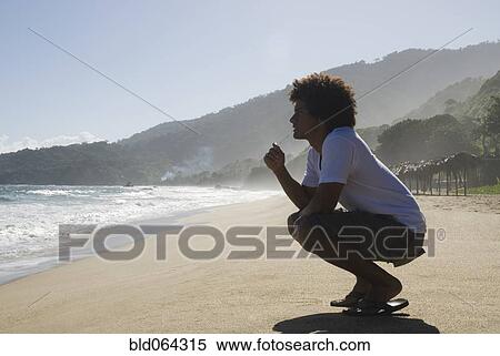 Stock Photography - African man crouching at beach. Fotosearch