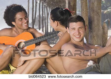 Stock Image - African man playing guitar for friends at beach. Fotosearch