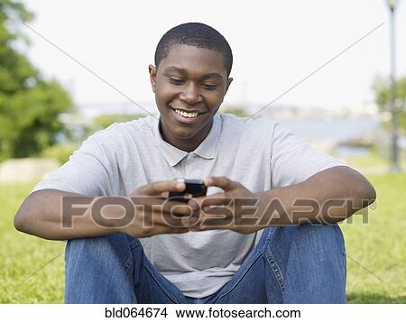 Picture - African man sitting in park text messaging on cell phone. Fotosearch