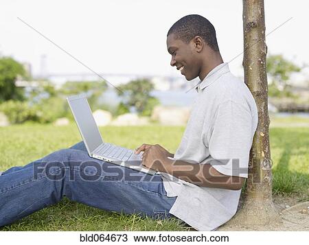 Stock Image - African man sitting in park typing on laptop. Fotosearch