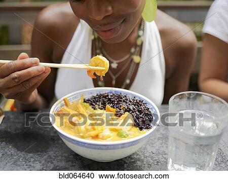 Stock Image - African woman eating Asian food in restaurant. Fotosearch