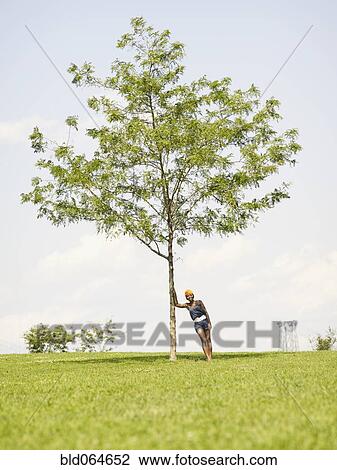 Stock Image - African woman leaning against tree in park. Fotosearch