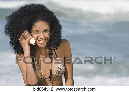 Stock Image - African woman listening to seashell on beach. Fotosearch