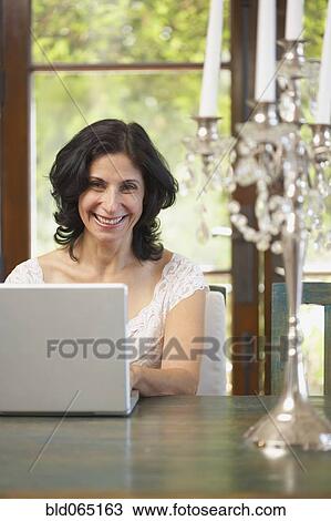 Stock Image - Confident Middle Eastern woman typing on laptop in dining room. Fotosearch