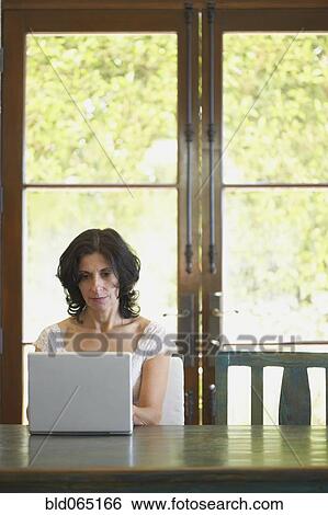 Stock Photograph - Confident Middle Eastern woman typing on laptop in dining room. Fotosearch