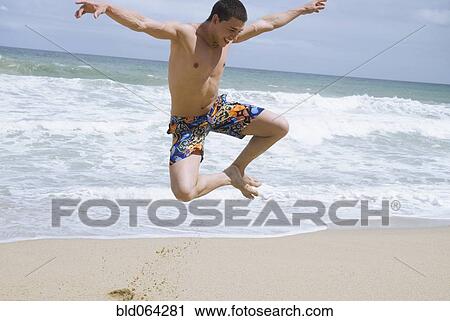 Stock Image - Hispanic man at beach jumping in mid-air. Fotosearch
