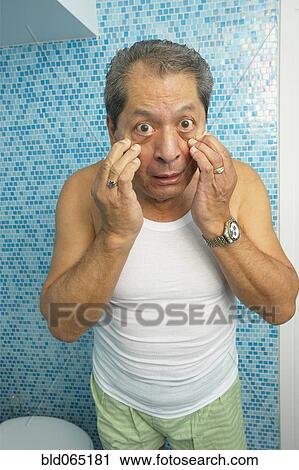 Stock Image - Hispanic man looking at his eyes in bathroom. Fotosearch