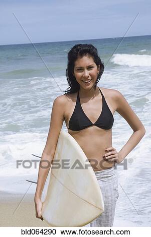 Stock Image - Hispanic woman holding surfboard at beach. Fotosearch