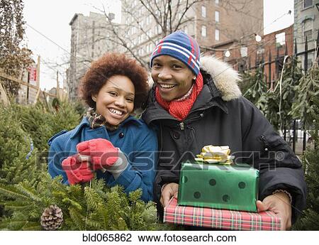 Stock Image - African couple in Christmas tree lot holding presents. Fotosearch