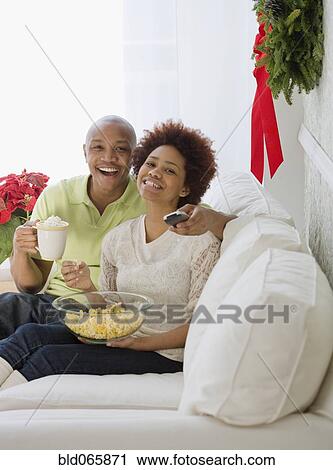 Stock Image - African couple watching television with popcorn and hot chocolate. Fotosearch