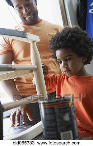 Stock Image - African father watching son paint rocking chair. Fotosearch