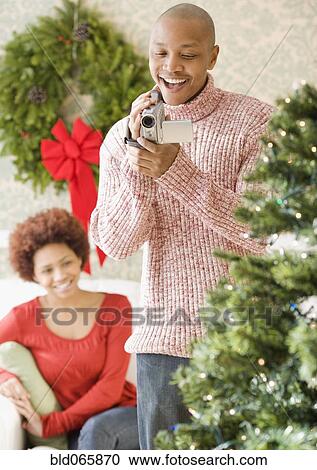 Stock Image - African man using video camera to film Christmas tree. Fotosearch