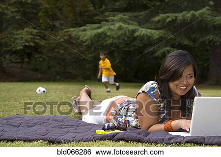 Stock Photograph - Asian mother using laptop in park with son in background. Fotosearch