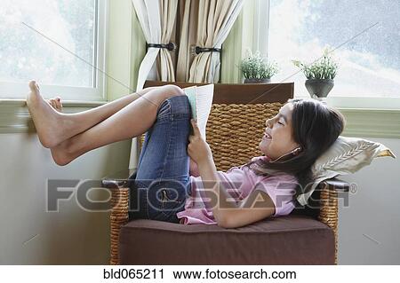 Stock Image - Hispanic girl reclining in armchair reading book. Fotosearch