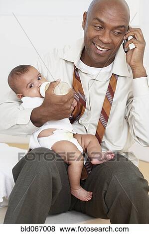 Stock Photo - African father feeding baby daughter and talking on cell phone. Fotosearch