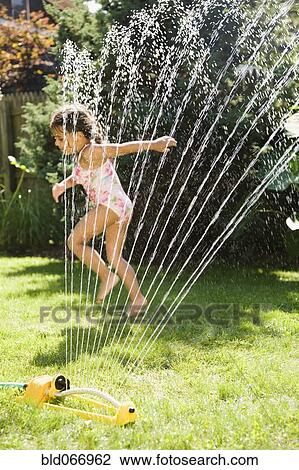 Stock Image - Hispanic girl playing in water sprinkler. Fotosearch