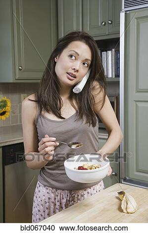 Stock Image - Woman eating cereal and talking on telephone in morning. Fotosearch