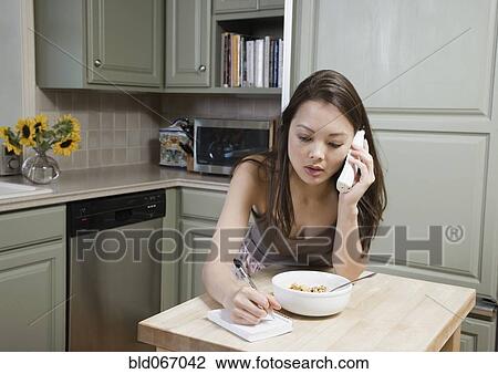 Stock Image - Woman eating cereal and talking on telephone in morning. Fotosearch