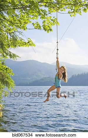 Picture - Asian girl hanging from lake rope swing. Fotosearch
