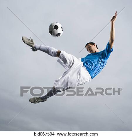 Picture - Mixed race teenager in mid-air kicking soccer ball. Fotosearch