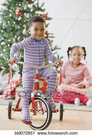 Stock Photo - African American boy riding tricycle near Christmas tree. Fotosearch