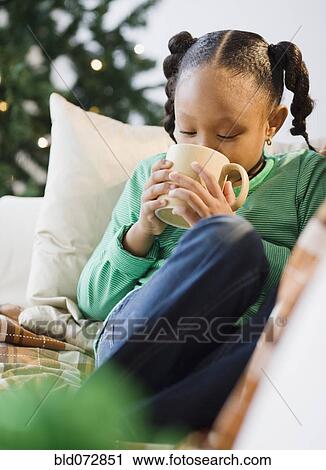 Stock Image - African American girl drinking hot chocolate on sofa. Fotosearch