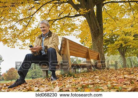 Stock Image - African man using cell phone on park bench in autumn. Fotosearch