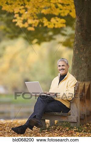 Stock Image - African man using laptop on park bench in autumn. Fotosearch