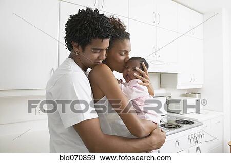 Stock Photo - African parents hugging baby in kitchen. Fotosearch