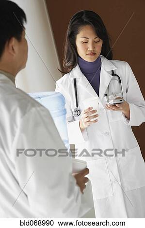 Stock Image - Asian female doctor using cell phone next to water cooler. Fotosearch